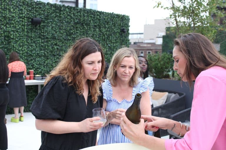 Three women engaged in a wine tasting event on a rooftop, attentively examining a bottle while holding glasses, with a lively backdrop.