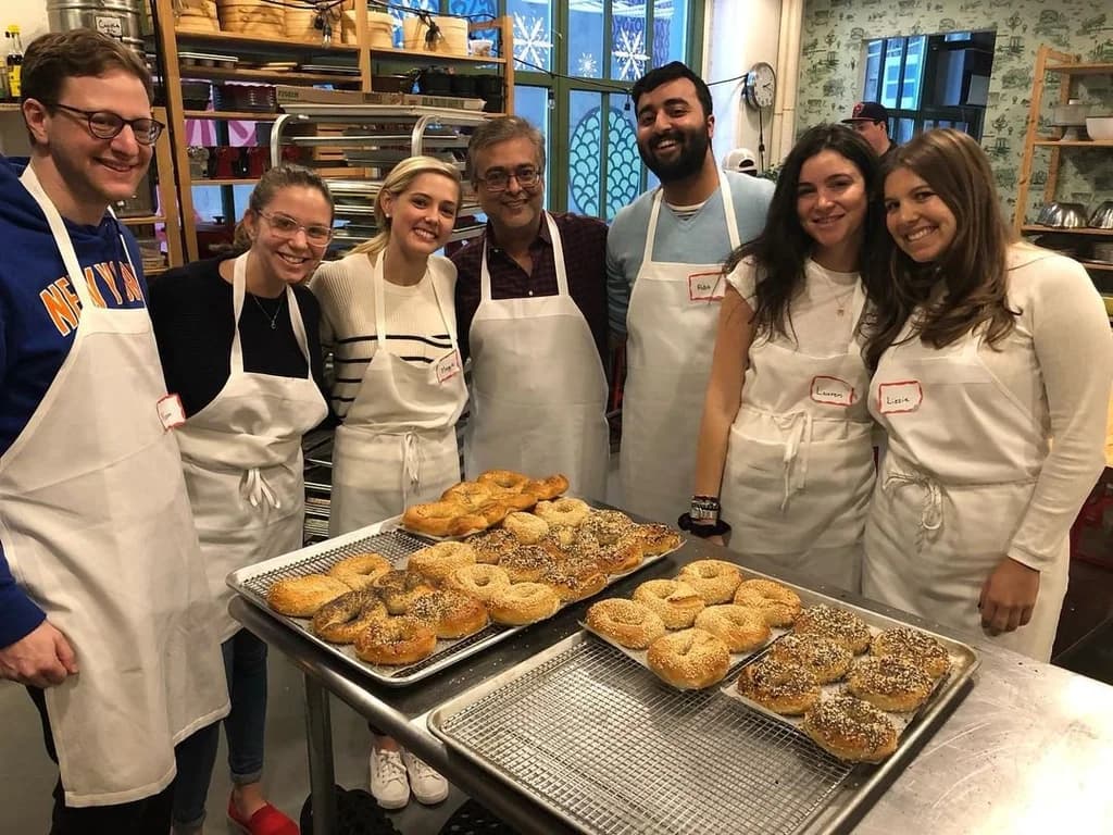 A group of people in aprons posing in front of trays of freshly baked bagels in a kitchen, smiling as they complete their baking class.