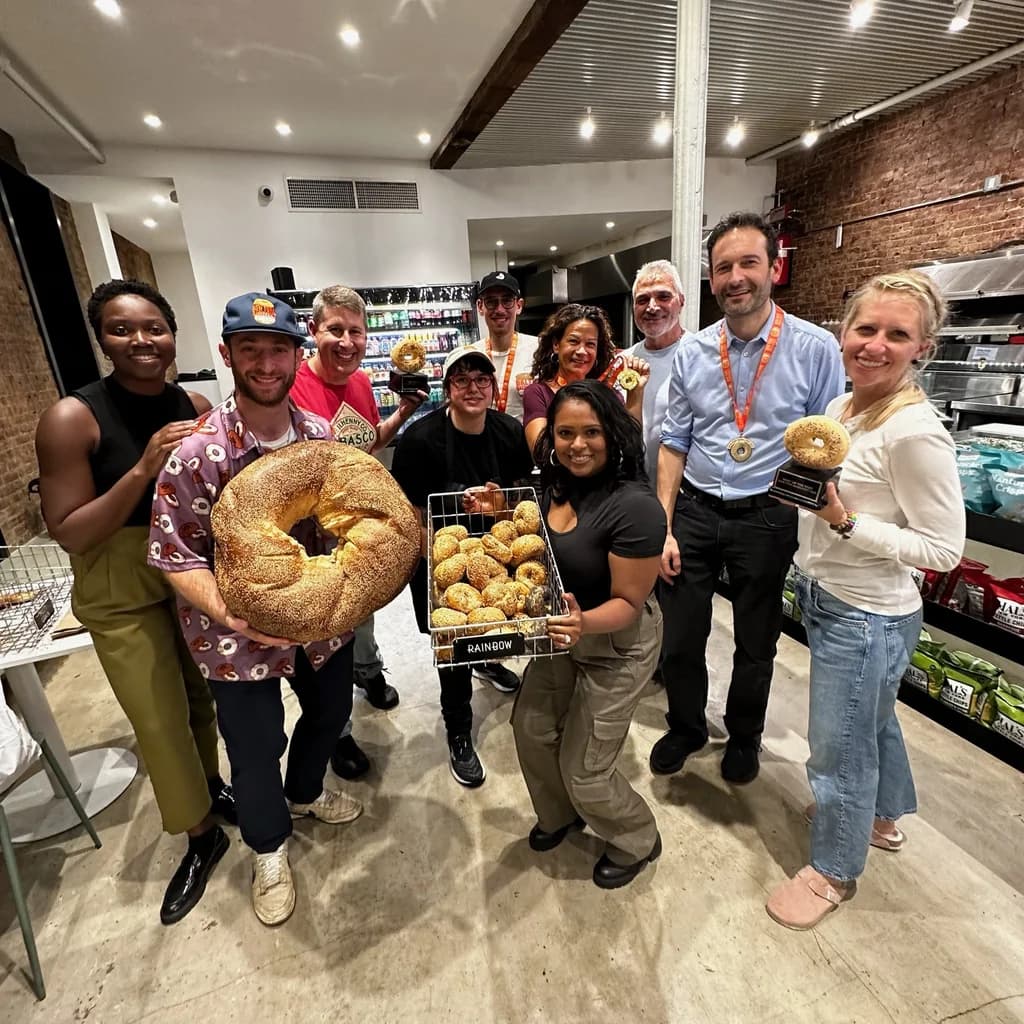 A group of people in a bakery proudly displaying an oversized sesame bagel and a tray of regular-sized bagels, smiling for the camera.