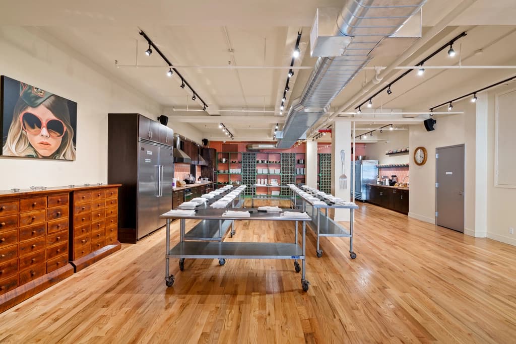 A well-lit kitchen studio with wooden floors, stainless steel tables, and cooking stations set up for a cooking class or event.