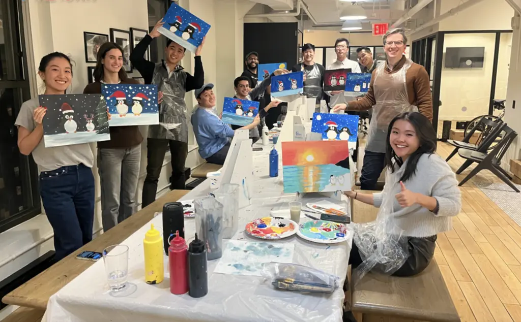 A group of people displaying their holiday-themed paintings at a painting workshop, with art supplies and easels on long tables.