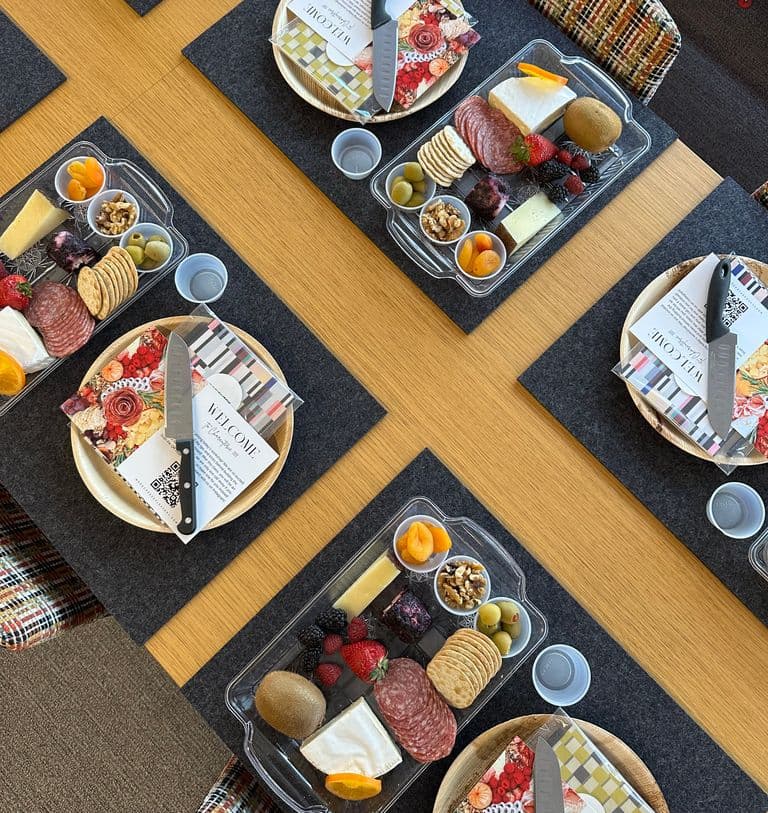 Overhead view of a conference table set with individual charcuterie trays, each featuring cheese, salami, crackers, fruit, and nuts, alongside a plate with napkin, welcome card, and knife on black placemats and colorful chairs.