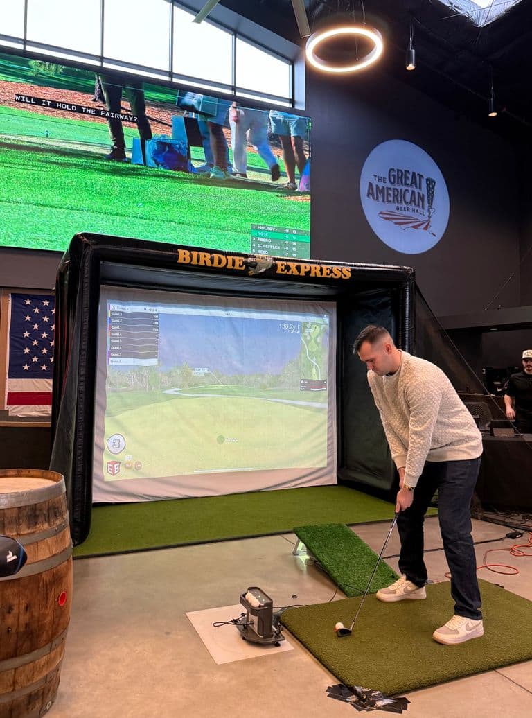 Person preparing to hit a golf ball in front of an indoor golf simulator setup, with a large screen displaying a live golf broadcast in a spacious venue.