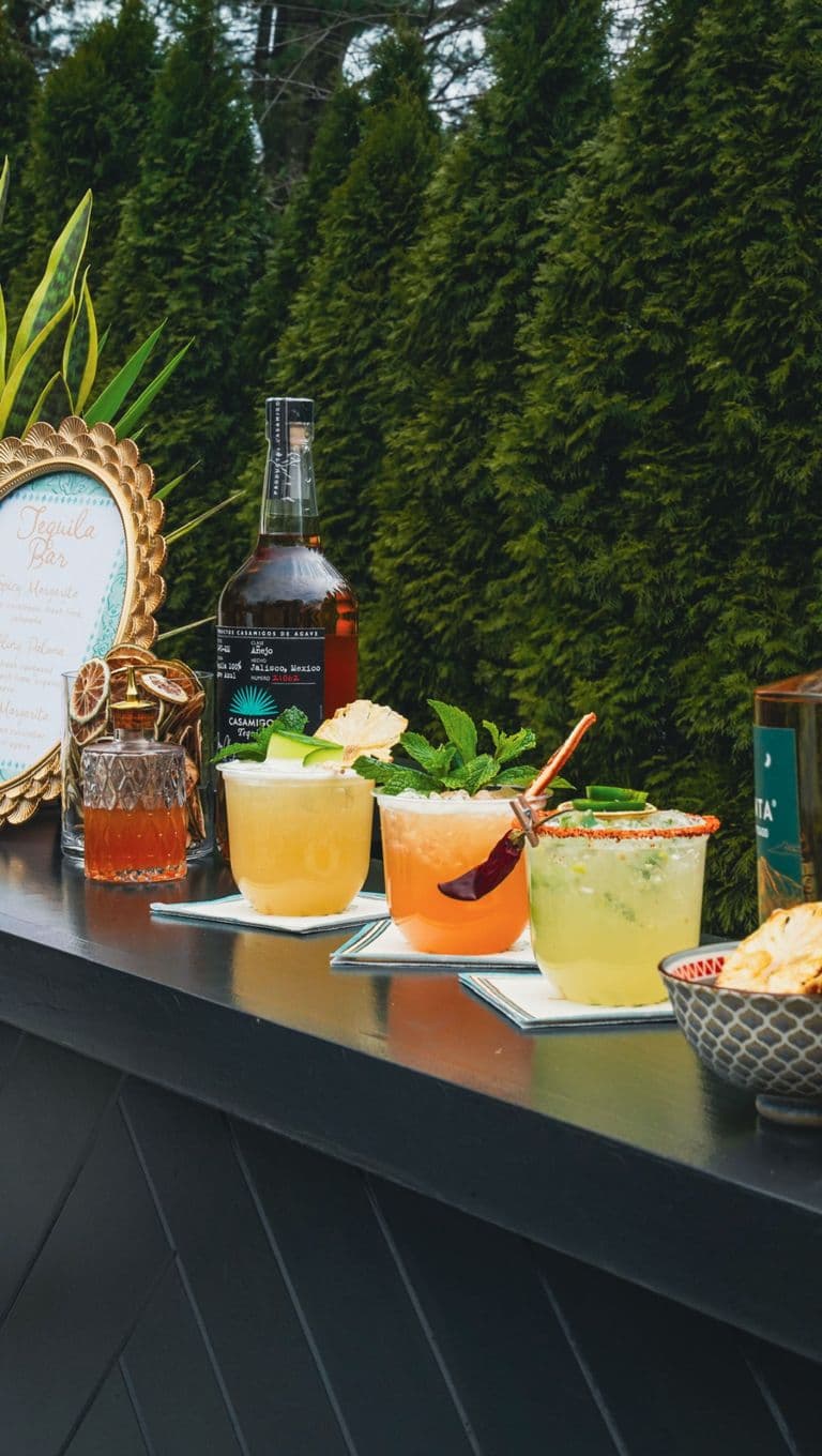 Close-up of a stylish outdoor tequila bar featuring a decorative drink menu, bottles of tequila, and four colorful cocktails garnished with fresh herbs, fruit, and chili, set against lush greenery.