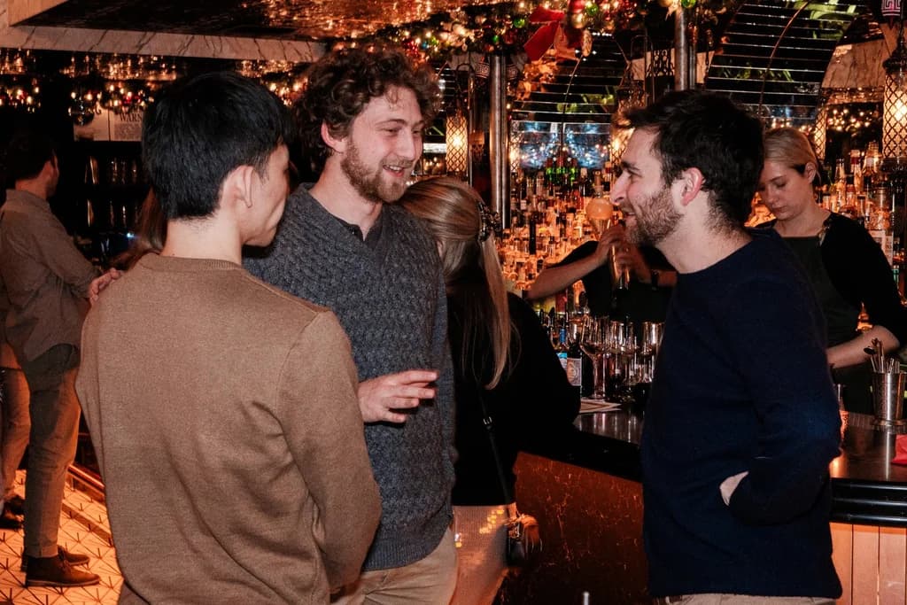 Three people engaged in a conversation at a bar, surrounded by festive lighting and a well-stocked counter.