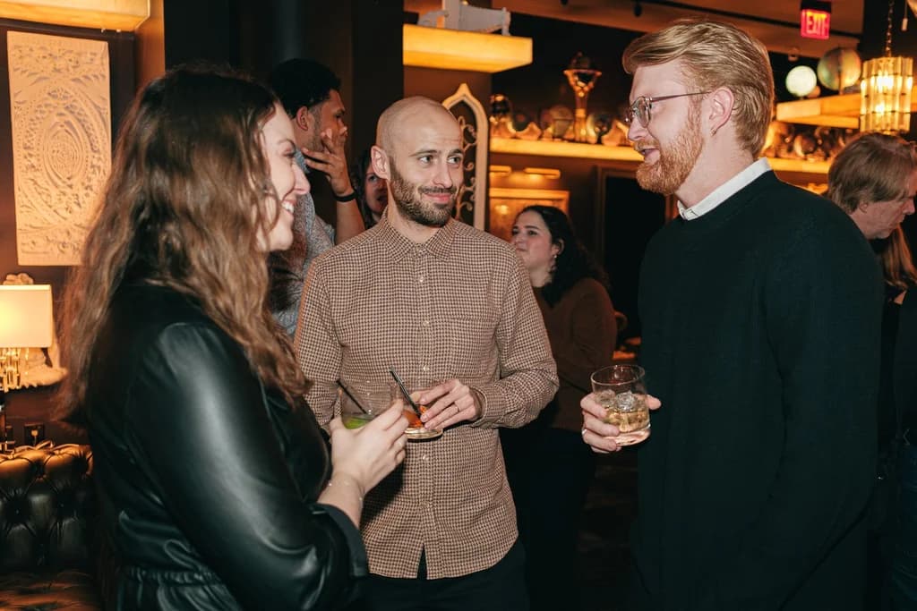 A group of people laughing and enjoying drinks at a cozy indoor gathering with warm ambient lighting.