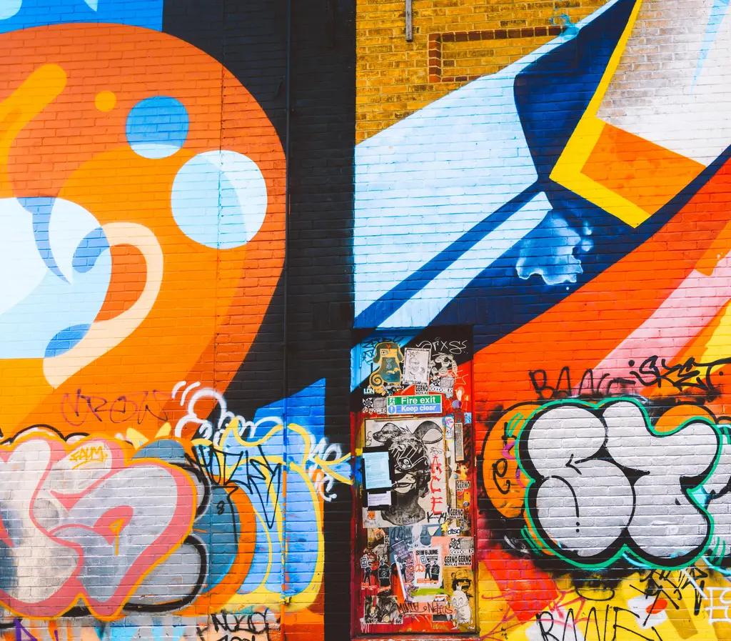 Colorful street art with bold geometric shapes, overlapping layers of graffiti, and stickers on a fire exit door on a brick wall.