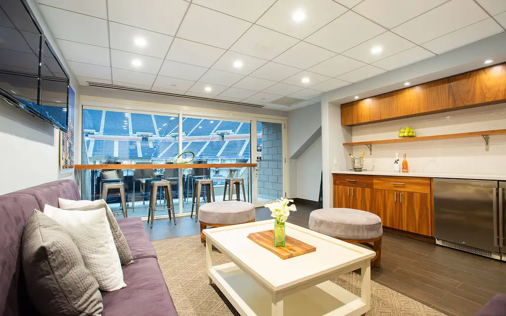 Private luxury suite with a view of the tennis court, featuring comfortable seating, a kitchenette, and bar stools for an elevated experience.
