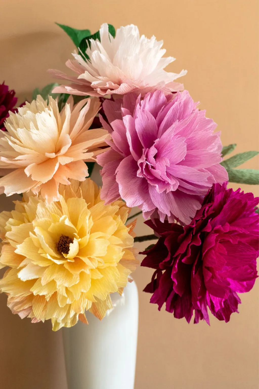 A beautiful bouquet of vibrant crepe paper flowers in shades of pink, peach, yellow, and burgundy displayed in a white vase against a warm backdrop.